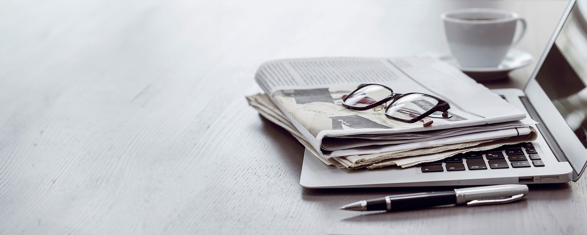 A laptop with a newspaper and glasses on top of it with a pen and a coffee mug on either side of it
