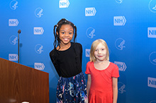 Two children are standing side by side and looking at the camera while smiling.