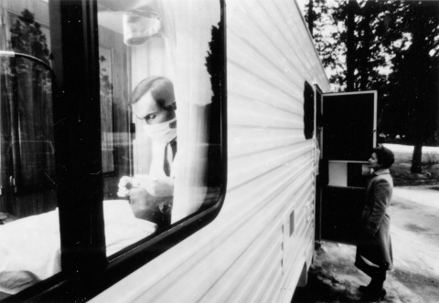 A dentist conducts a oral exam in an NIDR mobile dental examination unit and a woman stands outside waiting.