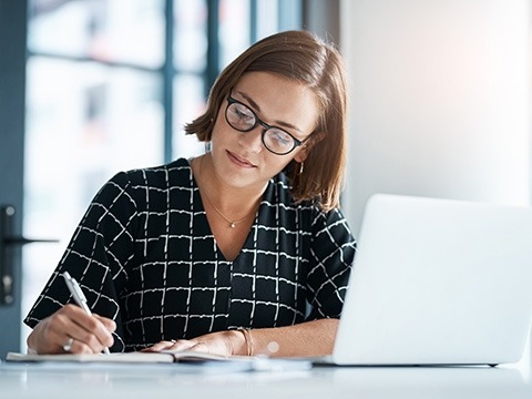 a women writing on a piece of paper next to a laptop