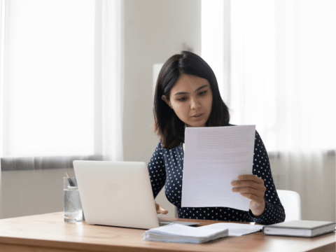 An image of a woman sitting at a desk, focused on a piece of paper in front of her.