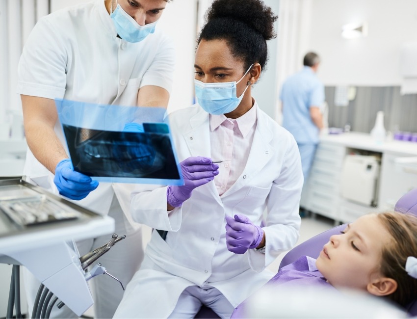 A clinician and dentist examine a radiograph in front of a patient seated in a dental chair in a dental clinic. The radiograph shows teeth and skeletal tissues surrounding the oral cavity.