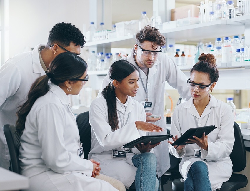 Three female researchers are sitting and two male researchers are standing above them and they are looking at a tablet