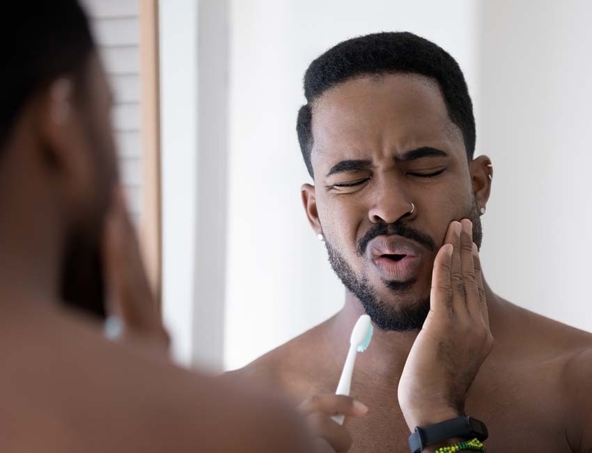 Hombre afroamericano cepillándose los dientes frente al espejo haciendo gesto de dolor dental. 