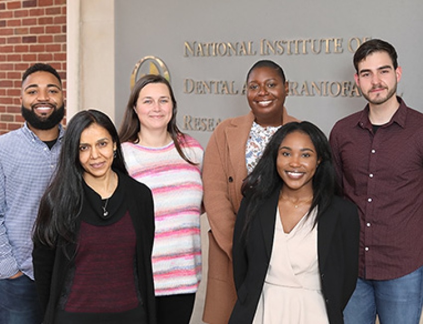 Six men and women standing together and smiling in front of a sign that says "National Institute of Dental and Craniofacial Research"