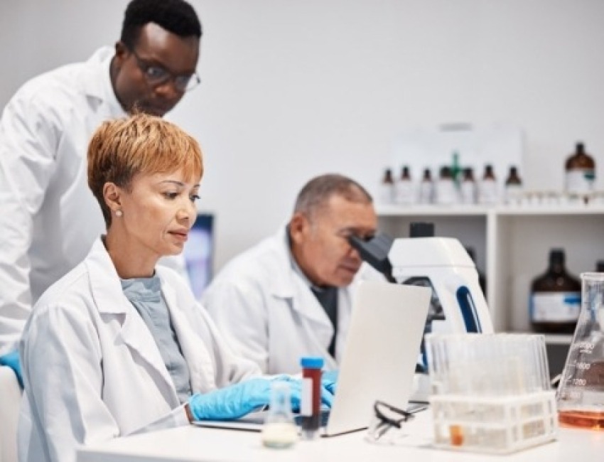 Female researcher looking at a laptop while a male researcher looks over her shoulder at the laptop and another male researcher sits next to her and looks into a microscope