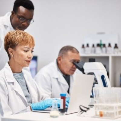 Female researcher looking at a laptop while a male researcher looks over her shoulder at the laptop and another male researcher sits next to her and looks into a microscope.