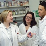 A group of three female and male scientists wearing lab coats and standing together in a laboratory. One of the female scientists stands in front of a white board holding a pen while gesturing and speaking to the other scientists.